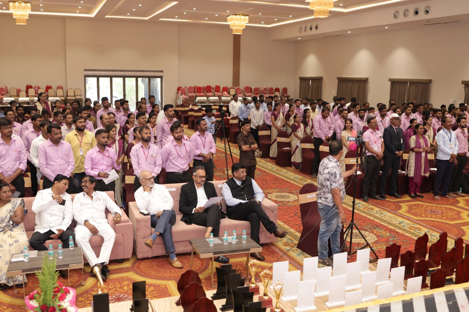Shivpratap Co-operative Credit Society staff in pink uniform shirts standing through an activity in the banquet hall, with the society’s office bearers seated in front, Karad