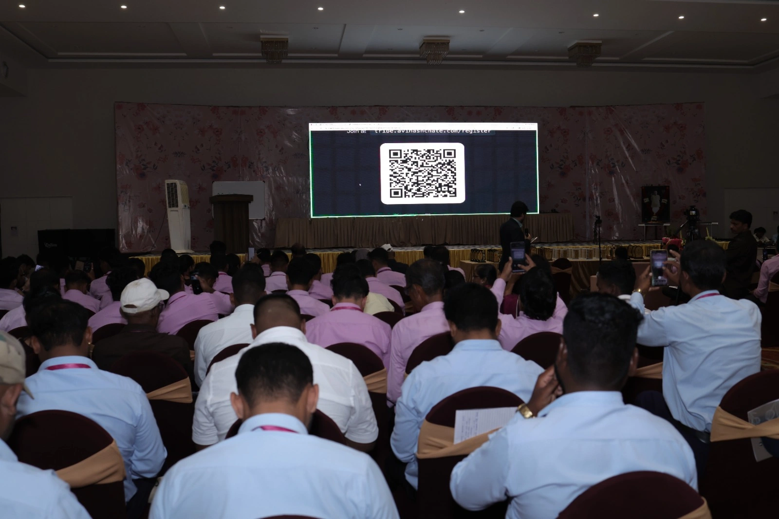 A full banquet hall of seated participants facing the stage as Avinash Chate presents, the LED screen behind him showing the session joining link