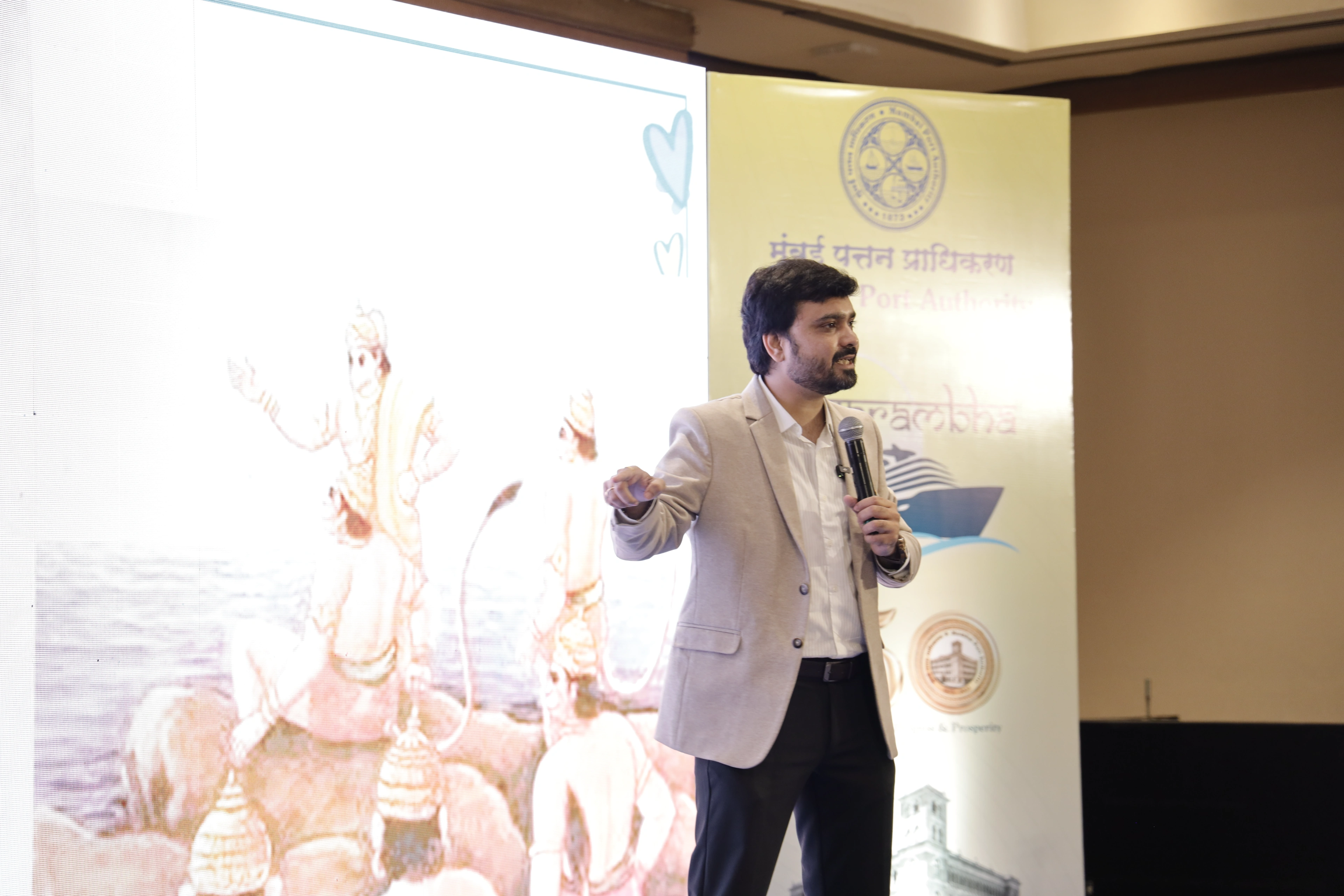Avinash Chate speaking with a microphone during the Mumbai Port Authority officers’ induction session, the authority’s Navarambha standee beside him