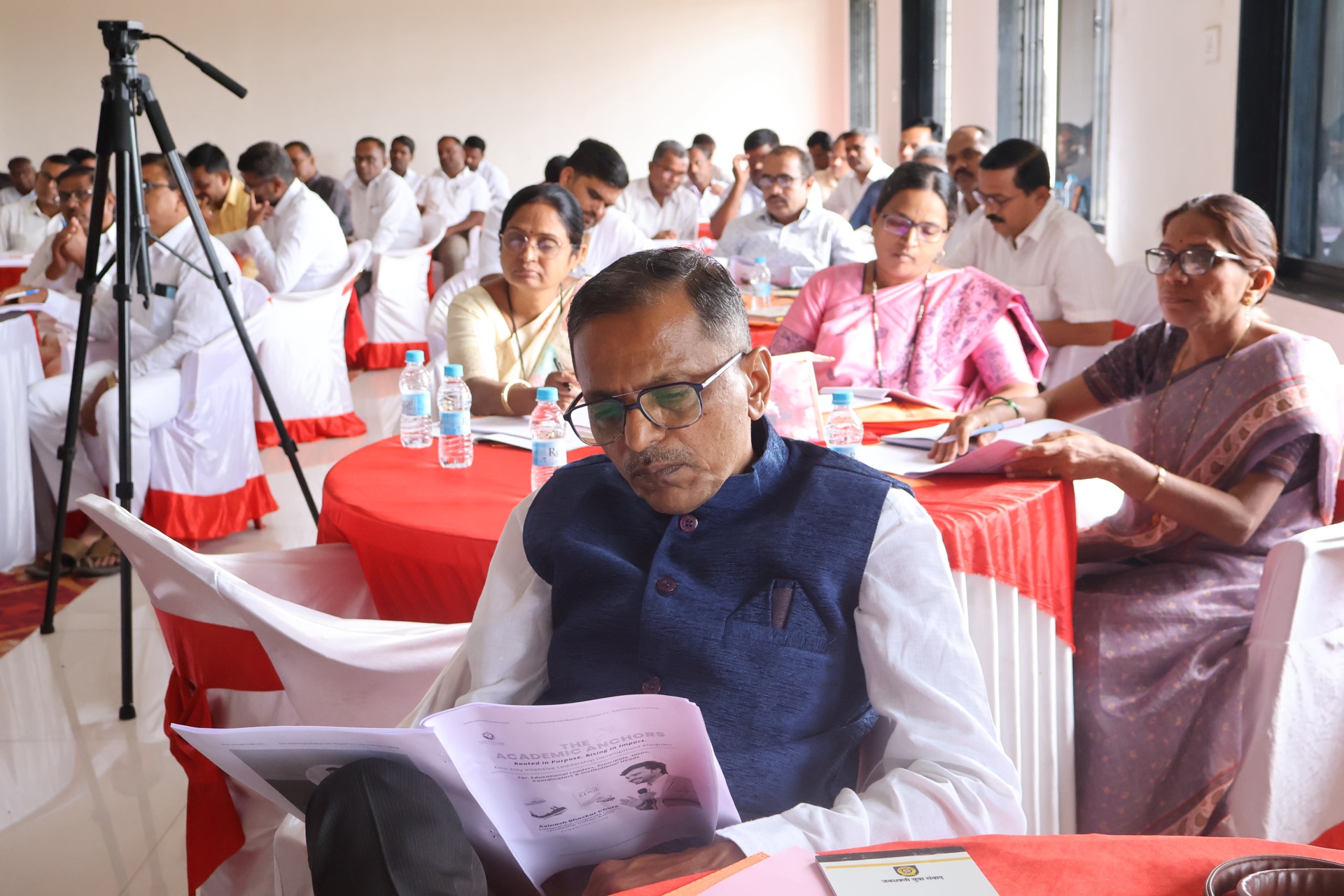 A school principal reading the printed programme booklet for the two-day leadership programme while fellow educational leaders take notes at round tables, Shevgaon